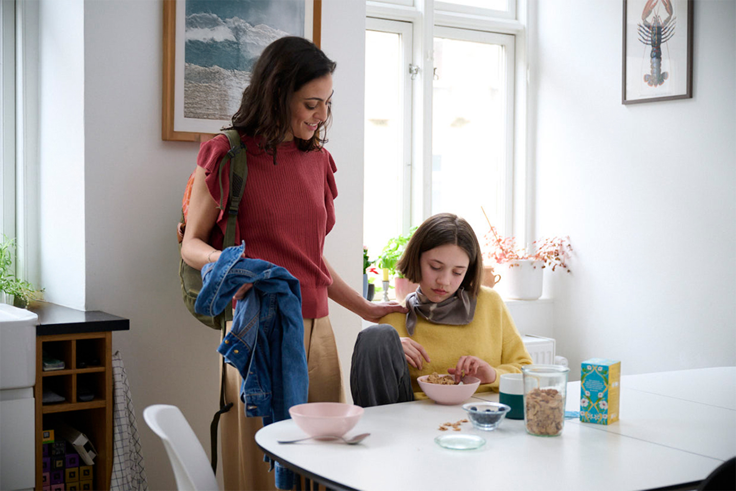 A woman rests her hand on the shoulder of a young girl looking sadly down at a bowl of food. 
