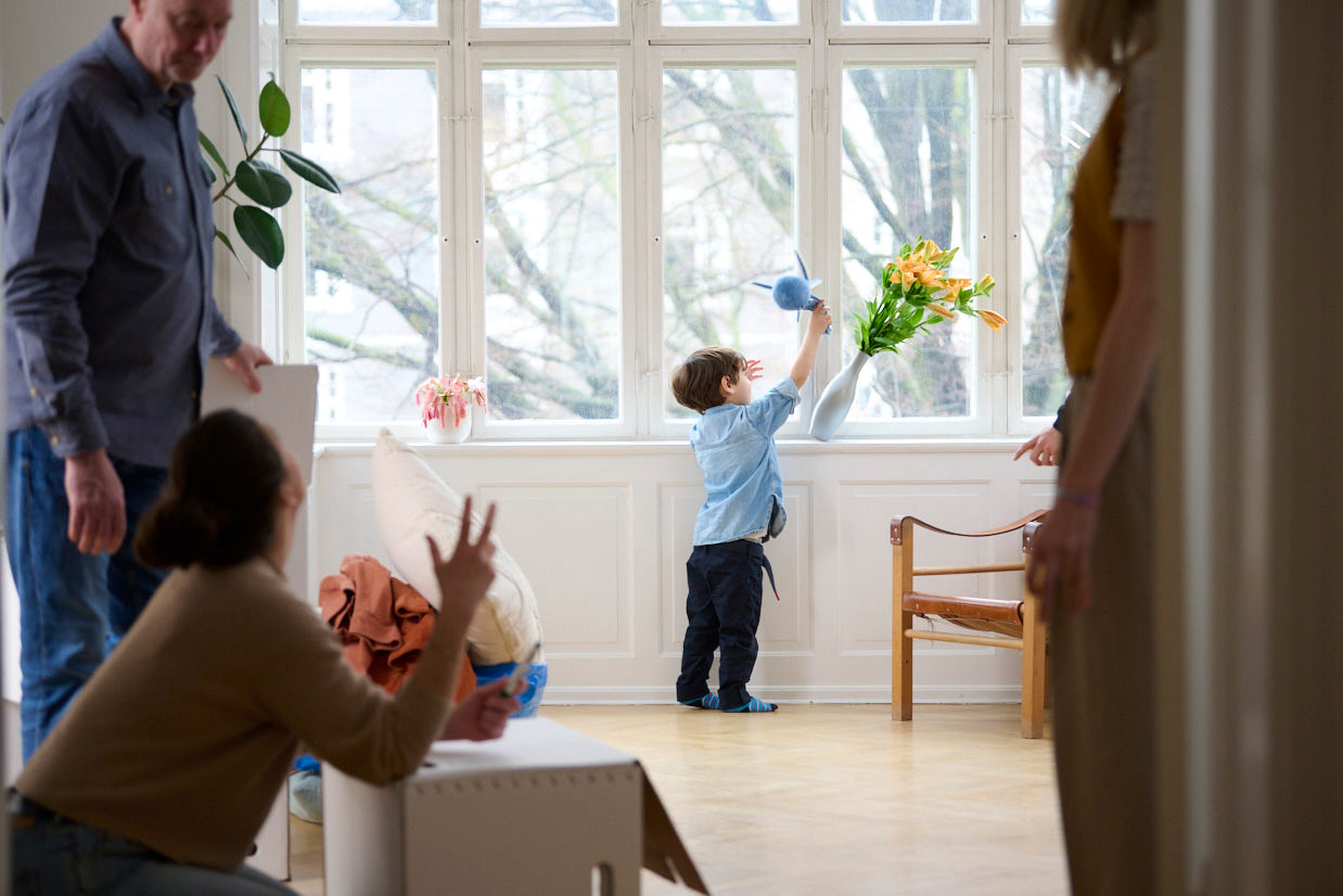 A child in the background knocks over a vase of flowers while two adults speak in the foreground